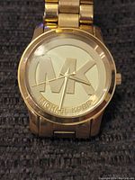 Front view of men's Michael Kors gold-tone watch showing large MK logo on the dial and metallic link bracelet.