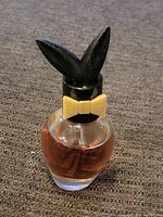Close-up of Playboy perfume glass bottle with black bunny ears cap, beige bowtie, containing brown liquid, approximately 70% full