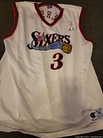 Front view of white basketball jersey featuring Philadelphia 76ers logo and number 3, with red and black collar and armhole trim.
