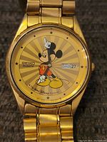 Close-up of watch face showing yellow dial with Mickey Mouse design, Seiko logo, and day/date display.