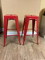 Pair of red metal bar stools standing on wooden floor against light brown wall, front view showing four legs and footrests