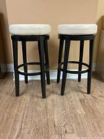 Photo showing two swivel bar stools side by side front view, featuring white round cushioned seats and black wooden legs on a hardwood floor against a beige wall.
