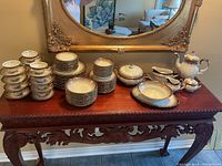 Full view of porcelain dinnerware set arranged on wooden table beneath ornate gold framed mirror. Includes teapot, sugar bowl, creamer, serving dishes, and multiple stacks of plates and cups.