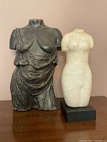 Two female torso busts, one dark grey with draped garment and one white on black base, placed on wooden surface against pink wall.