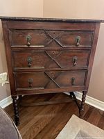 Front view of wooden cabinet showing three drawers with decorative carvings and metal handles, beaded trim on top, part of the turned legs and crossbars visible