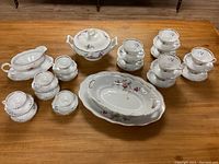 Full view of the multiple items arranged on wooden table, showing teacups with saucers, covered sugar bowl, gravy boat, large serving platter, and covered bowl.