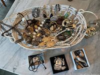 Wide view of white basket with assorted costume jewelry including necklaces, bracelets, earrings, and pins. Three boxes with additional jewelry next to the basket.