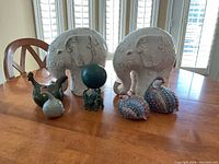 Photo displaying two large white elephant sculptures and smaller figurines including a green frog figurine holding a ball, a white bird figure, and a dark green bird figure on a wooden table.