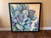 Framed square painting showing a close-up of a succulent plant with a black frame, leaning against a wall on wooden floor.