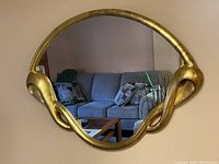 Front view of gold-toned oval mirror with artistic curved frame hung on beige wall above couch.