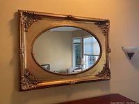 Frontal view of the large oval mirror with ornately decorated gold frame mounted on a wall.