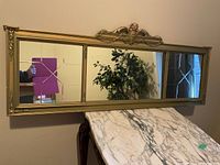 Full view of the large rectangular wall mirror mounted on the wall above a marble topped piece of furniture. The gold frame has an ornate central decorative crest and corner accents.