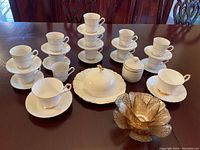 Various white ceramic teacups stacked on matching saucers, with a lidded sugar bowl and a covered butter dish also present. All pieces feature gold accent detailing and some signs of use such as crazing and scratches.