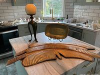 View of five carved wooden palm leaves laid out on kitchen island, next to green ceramic table lamp and rectangular ceramic tray on display stand.