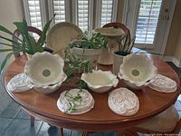 Photo of table displaying flower-shaped cream ceramic bowls, decorative ceramic plates, ceramic pots with living plants including trailing and succulent types in a well-lit room.