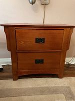 Front view of a medium-brown wooden side table with two drawers and black oval handles placed against a wall on a carpeted floor.