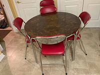 Round dining table with dark faux wood grain top surrounded by five red vinyl and chrome folding chairs on beige tiled floor