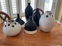 Five ceramic vases grouped on a wooden table with visible chairs and window blinds in the background. The vases are assorted in black and white colors, featuring multiple hole designs near the openings and varying shapes including tall and round.