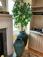 Two large floor vases placed beside a fireplace and cabinet — one smooth teal with dark handles, the other ribbed dark teal with artificial plant.