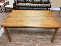Full view of the rectangular wooden table showing the table top and four legs, placed on a wood floor with chairs in the background.