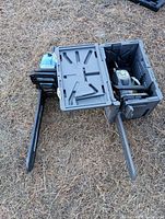 Two gas chainsaws placed side by side on the ground in open plastic carrying cases showing overall appearance of chainsaws and their cases.