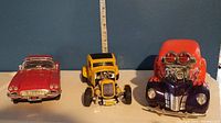 Front view of the three die cast cars with measuring tape behind for size reference. Showing red Corvette, yellow hot rod, and custom flame hot rod.