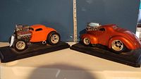 Image of orange Willys Coupe and metallic burnt orange Ford Coupe die cast muscle car models side by side on display bases with height ruler in background.