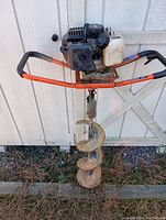 Front view of gas-powered post hole digger with orange frame, black engine, and white fuel tank propped against a shed wall.