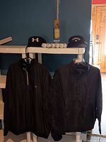 Two black golf jackets (Austin Reed XXL, Niklaus Large) and set of golf balls and visors on white shelf