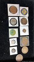 Coins and medals displayed in holders on a black felt background. Various sizes and designs, including Pope John Paul II labeled coin and colorful medallion.