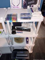 Wide view of the shelving unit holding various office supplies including a black IBM electric shredder on the bottom shelf, a black electric stapler and black three-hole punch on the middle shelf, and a large metal vintage trash can with floral design behind the shelving.