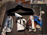 Lot showing two electric irons, white Steamfast garment steamer, 15 velvet hangers stacked behind, and white metal over-the-door hanger with rollers.