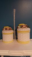 Two ceramic woven basket design jars with fruit and vegetable decoration on lids, placed side by side with a tape measure for scale.