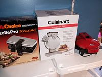 Photo showing three small appliances: Chef's Choice Pizzelle Pro Toscano Express Baker 834, Cuisinart Vertical Waffle Maker in box, and red Express Omelette Maker on white table.