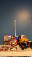 Overall image showing six toy cars including a yellow Brum character car and five packaged toy cars arranged in front of a blue wall and a measuring tape.