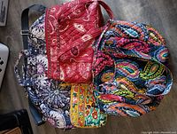 Four Vera Bradley bags arranged on floor showing variety of colors and patterns, including large duffel bag, two handbags, and small zip-up pouch.