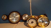 Three weather instruments including two polished brass and one wooden with multiple dials and barometers.
