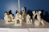 Overall view of collection of white textured Snowbabies porcelain figurines themed around Disney Snow White and the Seven Dwarfs on a white surface with a blue background wall and ruler indicating scale.