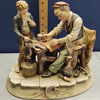 Porcelain figurine showing cobbler seated at workbench with boy standing beside him, measuring approx. 10 inches tall.