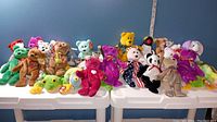 Wide image showing over 30 assorted TY Beanie Babies plush toys in multiple colors and species, placed on white tables against blue background.