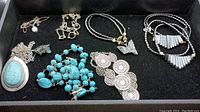 Top view of all jewelry in the lot placed in a black tray, showing turquoise pendant necklace, chunky turquoise bead necklace, crocheted circles, butterfly pendant necklace, and beaded fan pendant necklace.