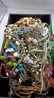 Photo of a black tray filled with a tangled assortment of costume jewelry featuring colorful beads, faux pearls, gold-tone and silver-tone chains, and various pendants.