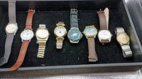 Full view of all eight men's watches arranged in a case showing varying strap materials and dial colors.