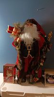 Full view of 24-inch rustic Santa figure with gift box ornament and another ornament box on white surface against blue wall