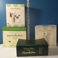 Three original boxes for Snowbabies figurines stacked together against a blue background with measuring tape showing scale.
