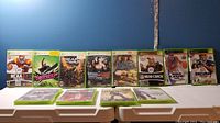 Xbox 360 game cases lined up on a table showing titles including NCAA 07, Amped 3, Gears of War, Smackdown VS Raw 2010, Blazing Angels, The Bigs 2, NBA 2K16, DEFJAM Rapstar, Dead Space 3, NFL Head Coach, NBA Live 2005, NCAA 05