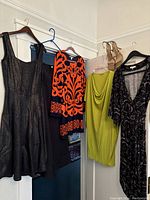 Four dresses hanging to display styles and colors: black sleeveless, orange/black patterned, lime green draped, black multicolor pattern dress