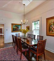 Full view of mission style dining table with 10 chairs around, set in a decorated room with paintings and chandelier overhead.