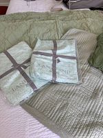 Two new packaged Sanderson top sheets folded and tied with ribbon on a bed, next to fitted sheet and shams.