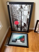 Two framed artworks, one large stretched canvas with black frame showing a black and white street scene with a red umbrella, and one smaller framed print laying on the floor.